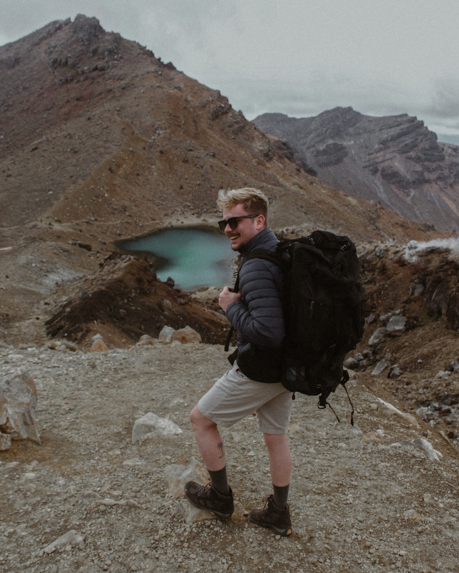 man in black jacket and white pants standing on rocky ground near lake during daytime