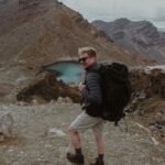 man in black jacket and white pants standing on rocky ground near lake during daytime